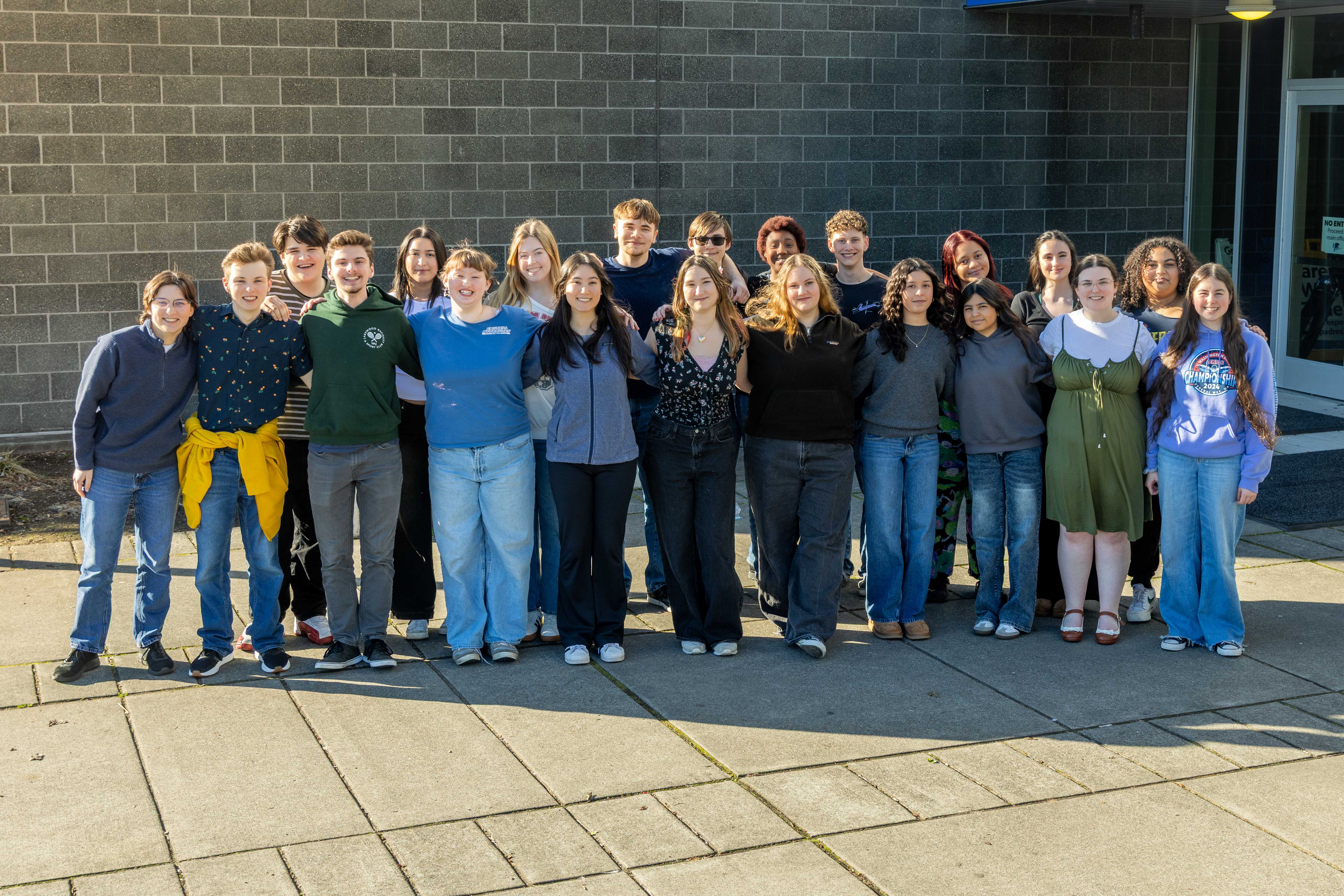 Harrison Preparatory School drama students standing in two rows while smiling with their hands over one anotherundefineds shoulders in front of a brick wall outside of the school.